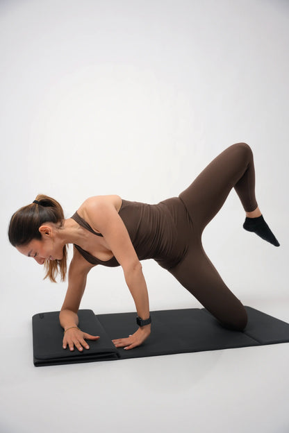 Woman in brown outfit performing a yoga pose on a black mat against a white background